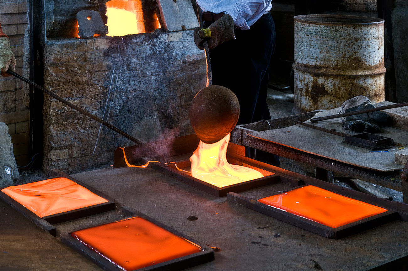 Gros plan sur un verrier versant du verre en fusion incandescant à l'aide d'une louche métallique dans des moules rectangulaires pour former d'épaisses dalles de verre.