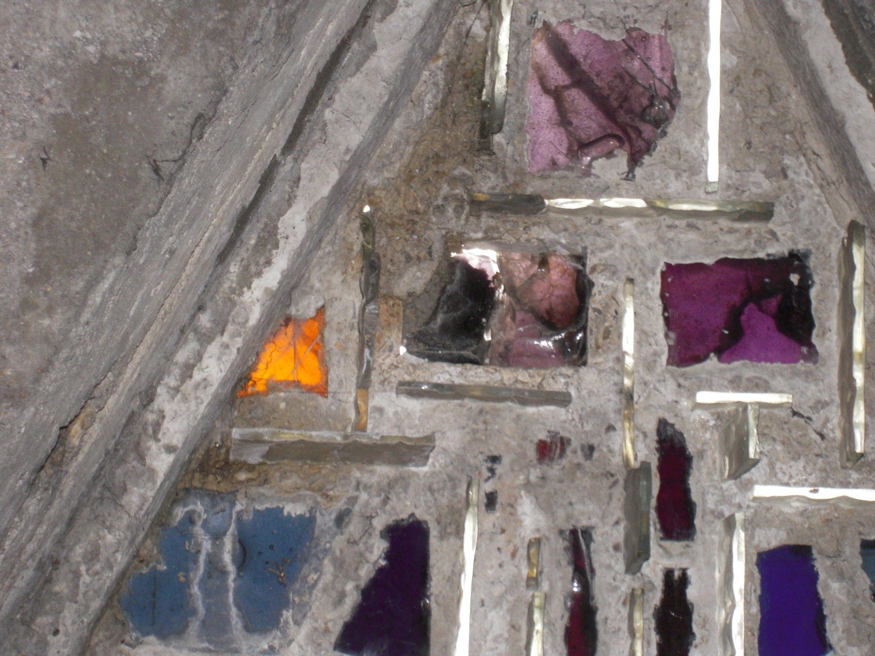 Gros plan sur la section triangulaire supérieure d'une baie en béton ou en pierre, montrant une structure en ciment brut encastrant des dalles de verre taillées aux teintes violettes, bleues et ambre.
