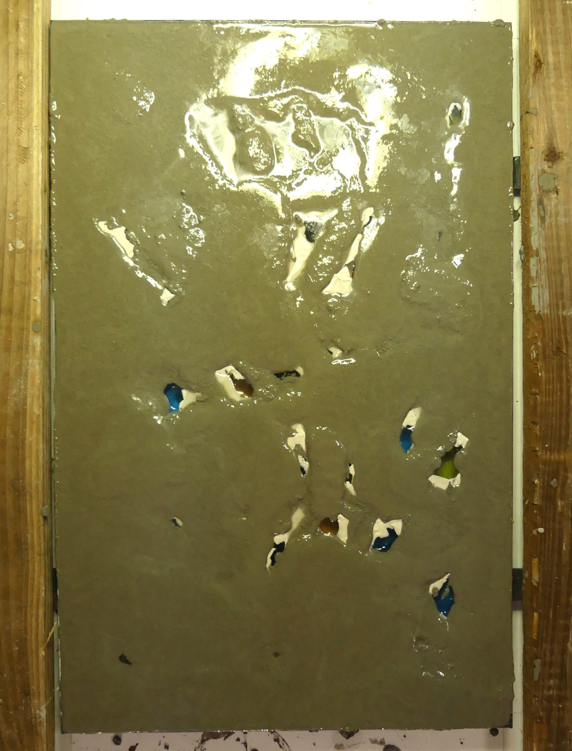 Panneau rectangulaire en cours de coulage montrant une structure en ciment encore humide recouvrant la surface, d'où émergent partiellement des éclats de dalles de verre colorées.