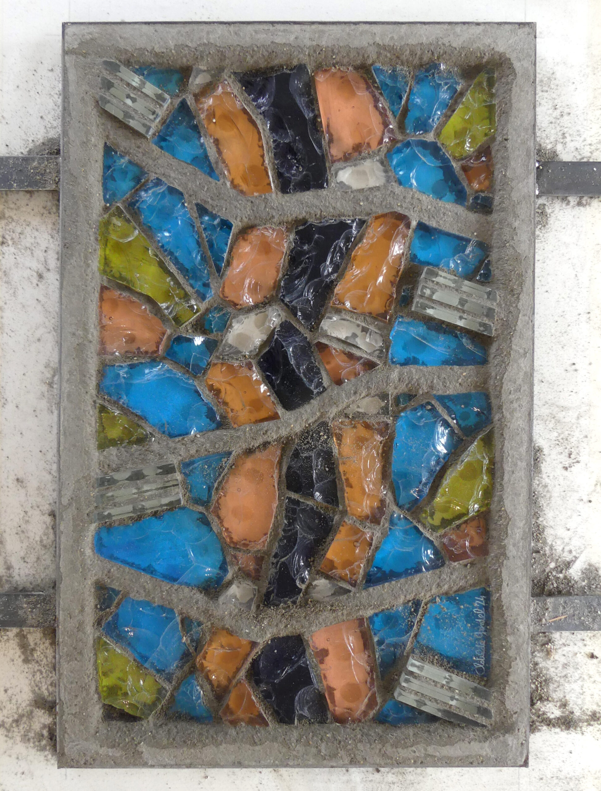 Panneau finalisé de dalle de verre rectangulaire, enserré dans une structure en ciment gris séché qui scelle solidement les morceaux de verre taillés bleus, ambre, verts, roses et translucides.