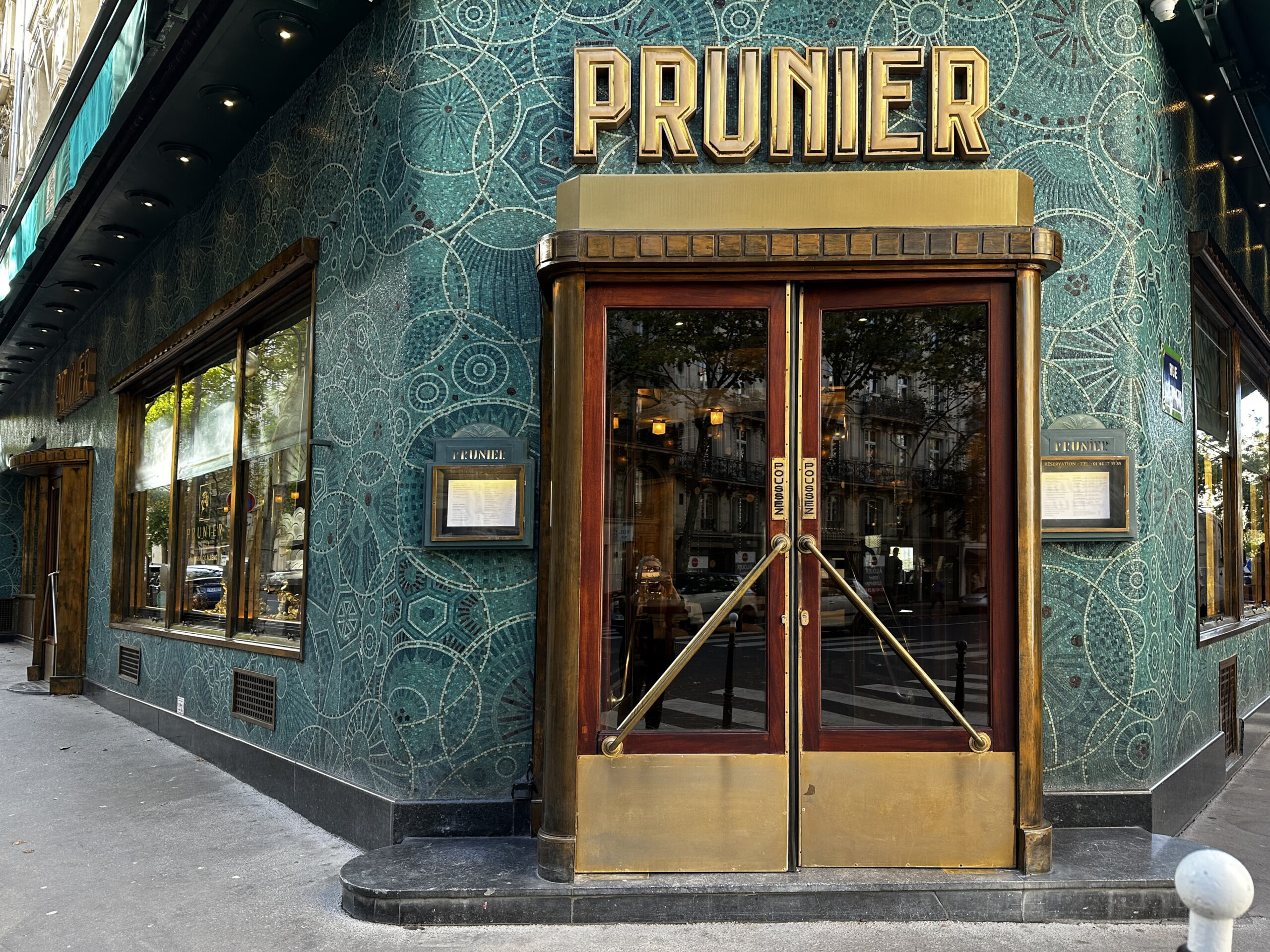 Façade de style Art déco du restaurant Prunier, recouverte de mosaïques vert turquoise à motifs circulaires et surmontée d'une enseigne dorée au-dessus de portes en bois et en laiton.