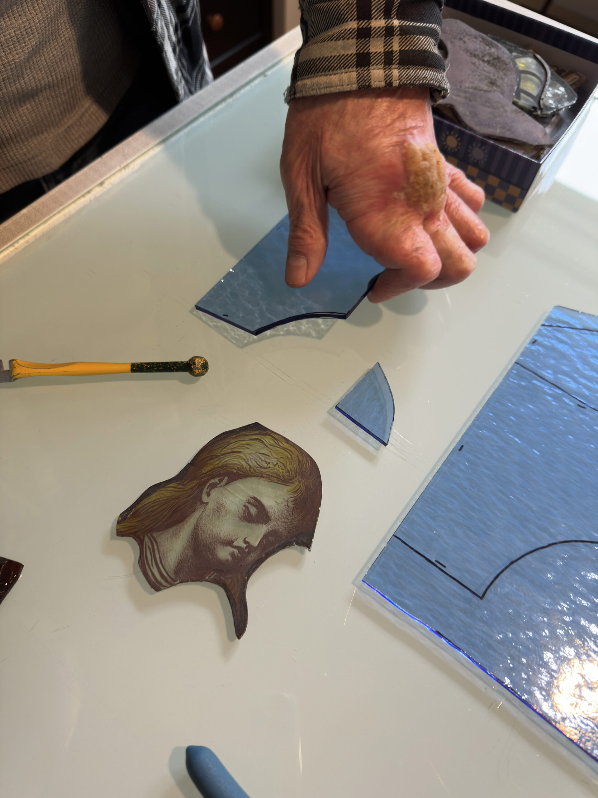 Gros plan sur la main d'un maître verrier ajustant des pièces de verre découpées et un fragment peint du visage d'un personnage sur une table lumineuse d'atelier.