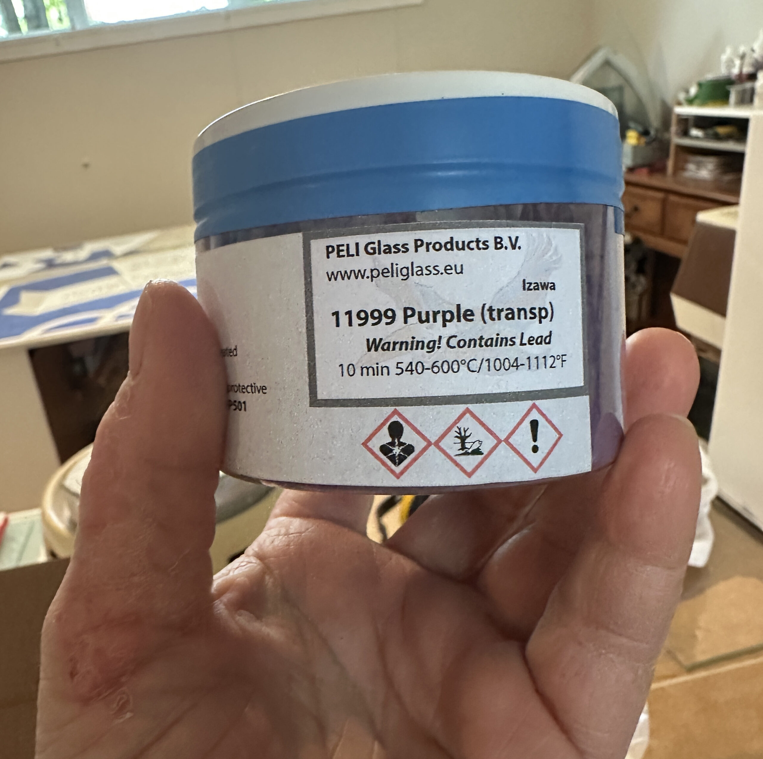 Gros plan sur une main tenant un pot de pigment pour verre de la marque PELI Glass Products, portant l'étiquette « 11999 Purple (transp) » avec des avertissements sur la présence de plomb et les températures de cuisson.