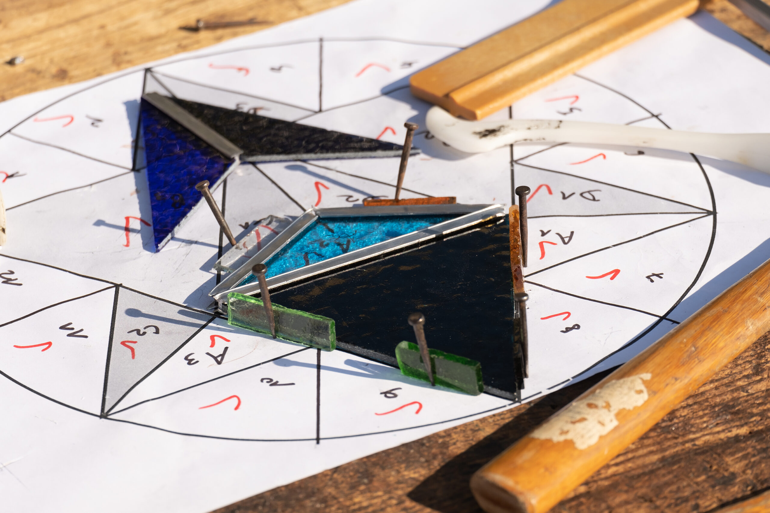 Vue sur un plan d'assemblage technique de vitrail géométrique, présentant des pièces de verre triangulaires bleues et sombres maintenues par des clous sur le tracé numéroté.