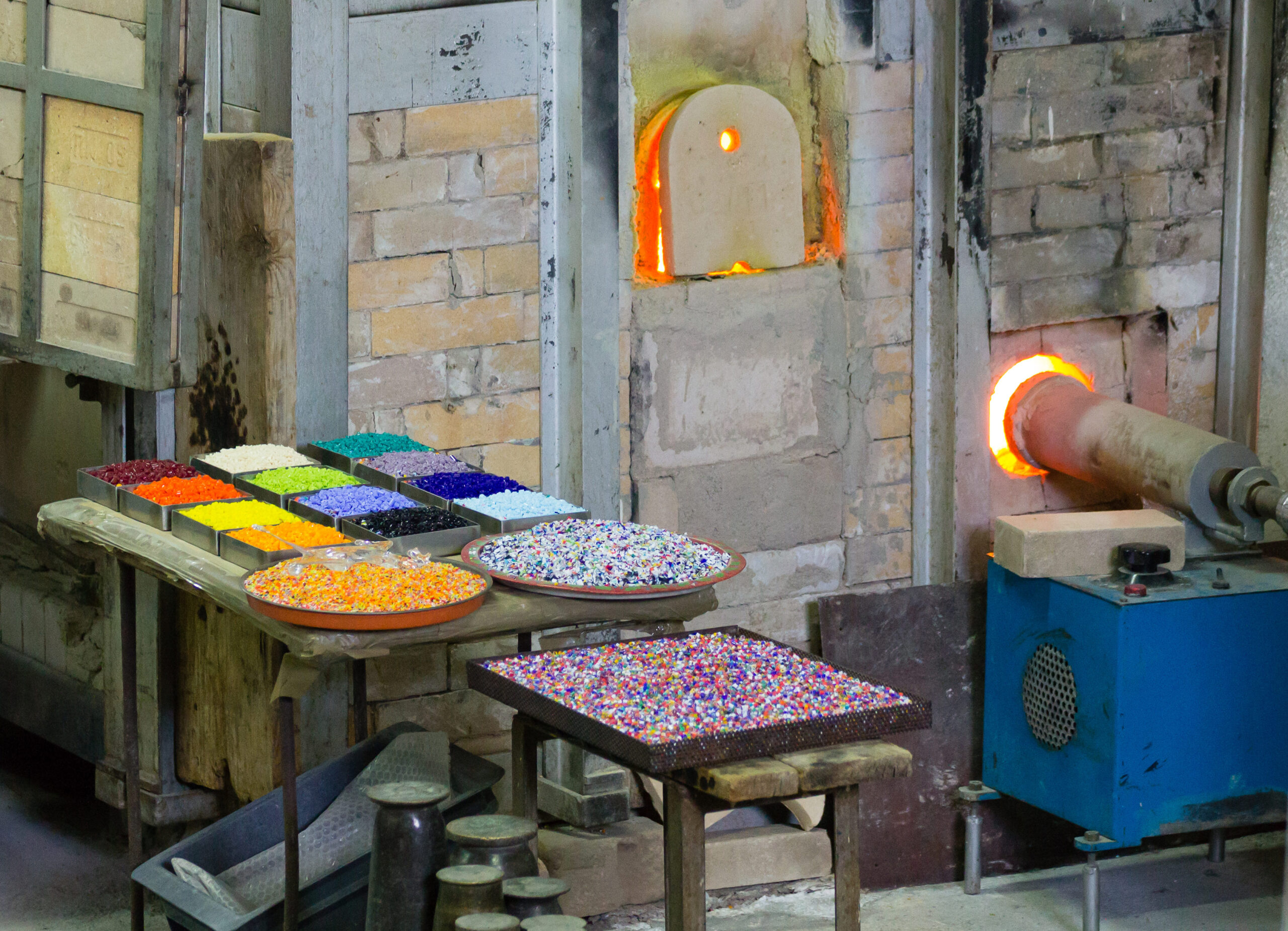 Vue d'un atelier de verrerie montrant des fours à haute température incandescents et une table garnie de récipients contenant des granules de verre de différentes couleurs.
