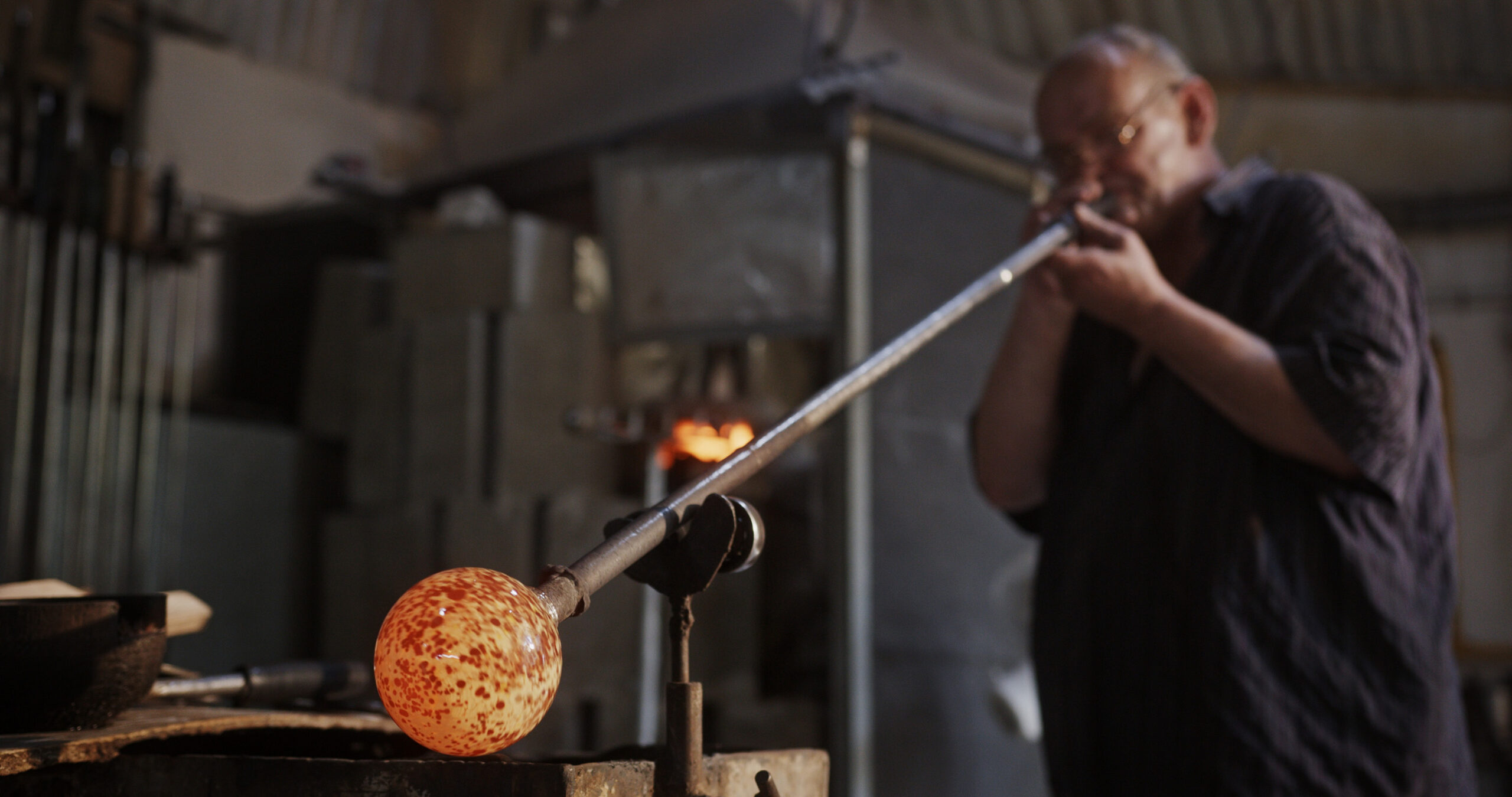 Un artisan souffleur de verre souffle dans sa canne pour façonner une sphère de verre incandescente aux teintes chaudes et piquées dans un atelier sombre.