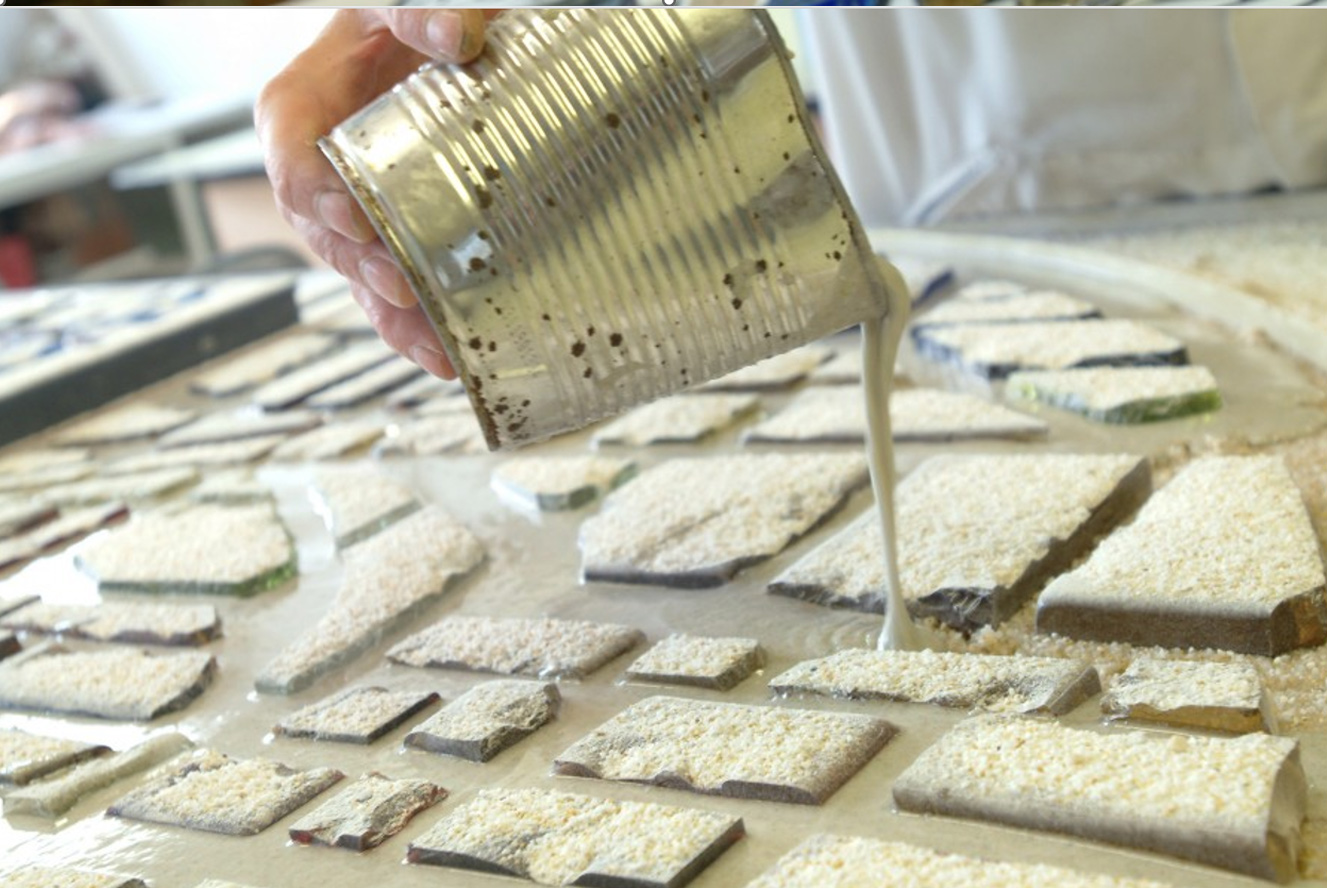 Gros plan sur la main d'un artisan versant un liant liquide depuis une boîte métallique entre des dalles de verre taillées disposées à plat sur un lit de sable pour former la structure en époxy.