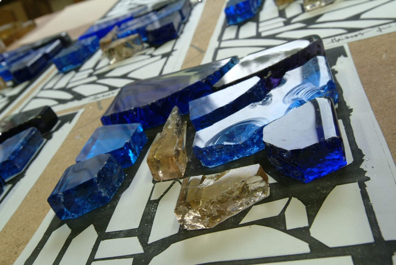 Vue en gros plan des dalles de verre taillées aux reflets bleus et ambre, aux bords écaillés, disposées sur un carton de travail tracé de lignes sombres.