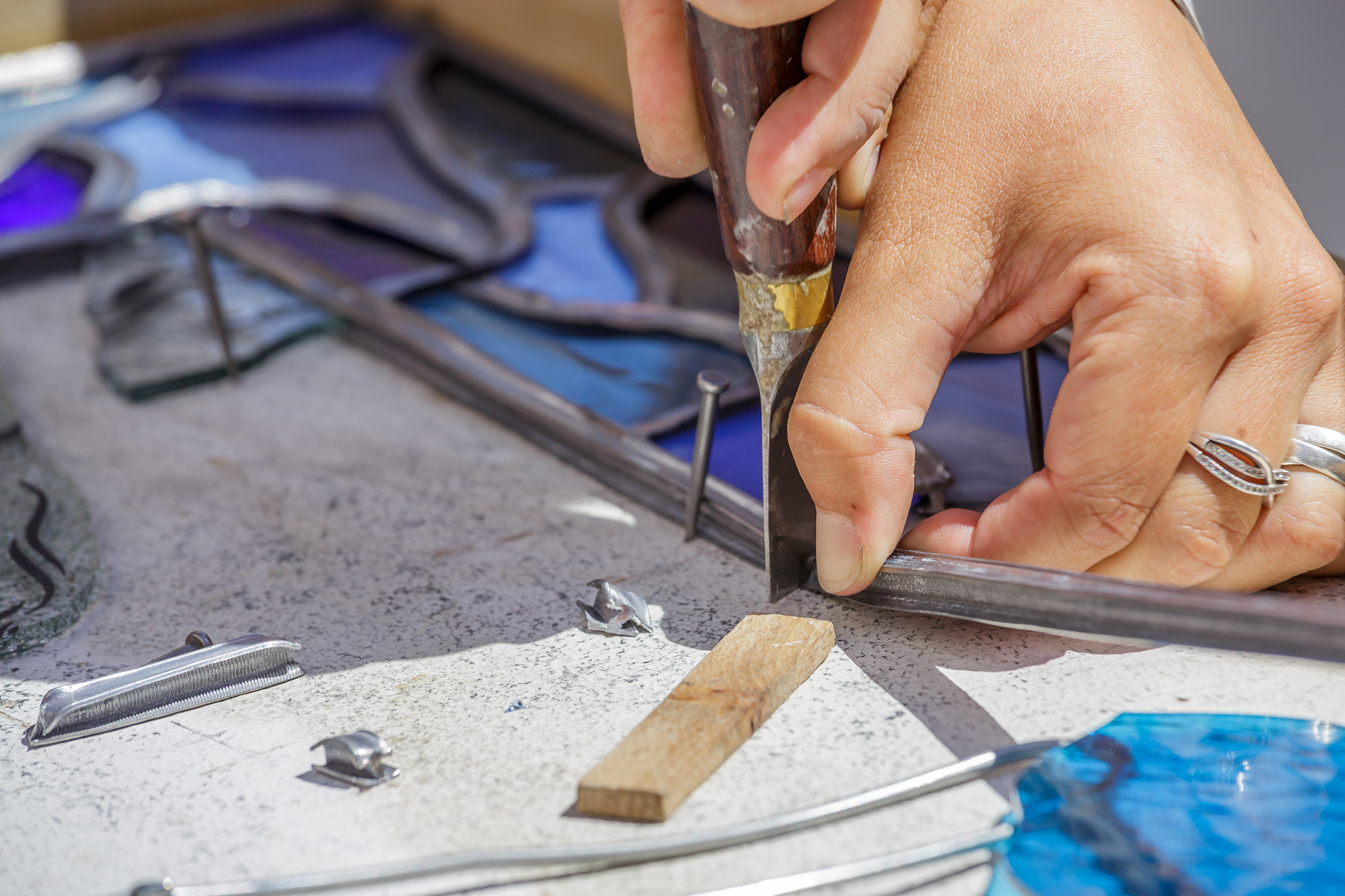 Gros plan sur les mains d'un artisan utilisant un couteau à plomb pour découper avec précision une baguette de plomb lors de l'assemblage d'un vitrail.